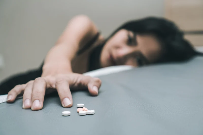 Young sad woman struggling with depression and prescription pill addiction reaches for a pile of pills